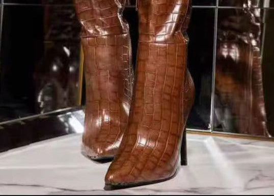 Brown crocodile-patterned knee-high boots worn by a person against a dark background.